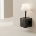 A black smart wall plug lamp with a pleated white mini lampshade and a built-in European wall outlet, shown plugged into a wall in a minimalist room setting.