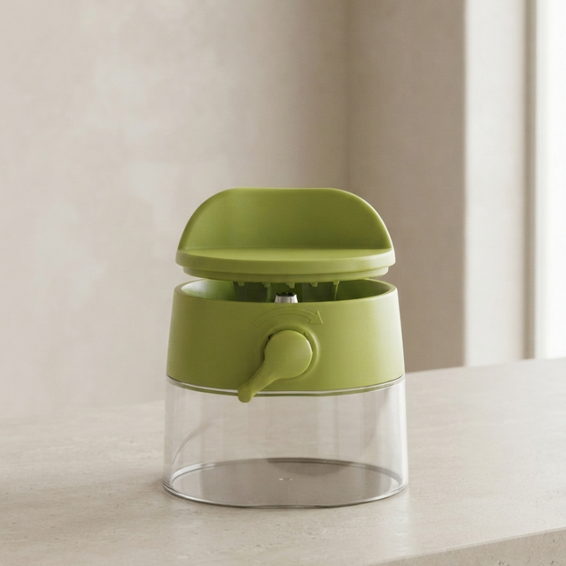 A small kitchen dispenser with a translucent clear body, a light olive-green plastic top and handle, and a matching green central collar, shown on a light beige countertop against a soft beige wall.