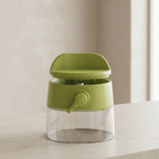 A small kitchen dispenser with a translucent clear body, a light olive-green plastic top and handle, and a matching green central collar, shown on a light beige countertop against a soft beige wall.