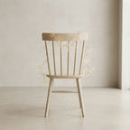 A light natural wood spindle-back chair centered in a minimalist room, decorated with delicate warm white fairy lights draped around the backrest.