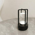 A modern black lantern with a clear glass cylinder and a glowing white interior, set on a gray marble surface against a neutral beige background.