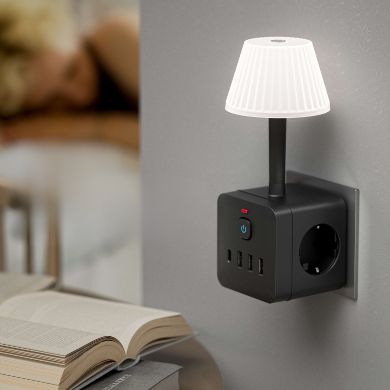 A black wall-plugged power strip night light with a small white pleated lampshade and multiple USB charging ports, shown mounted on a gray wall in a bedroom setting with an open book in the foreground.
