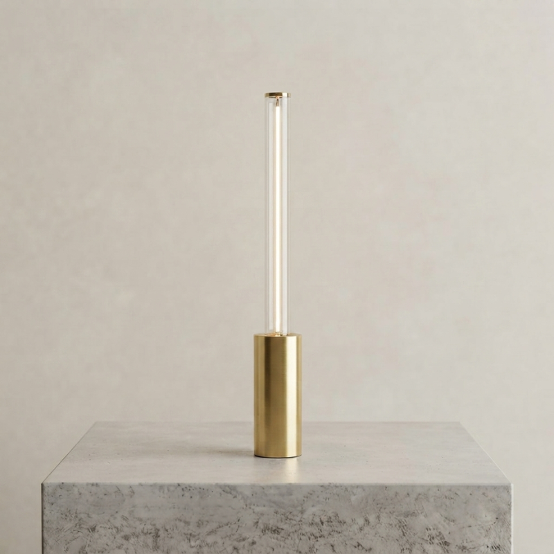 A modern table lamp with a cylindrical gold base and a clear glass tube, shown on a light gray stone pedestal against a neutral beige background.