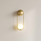 A modern wall-mounted light fixture with a brushed gold finish, featuring a spherical top and a glowing oval bottom, set against a plain off-white wall.