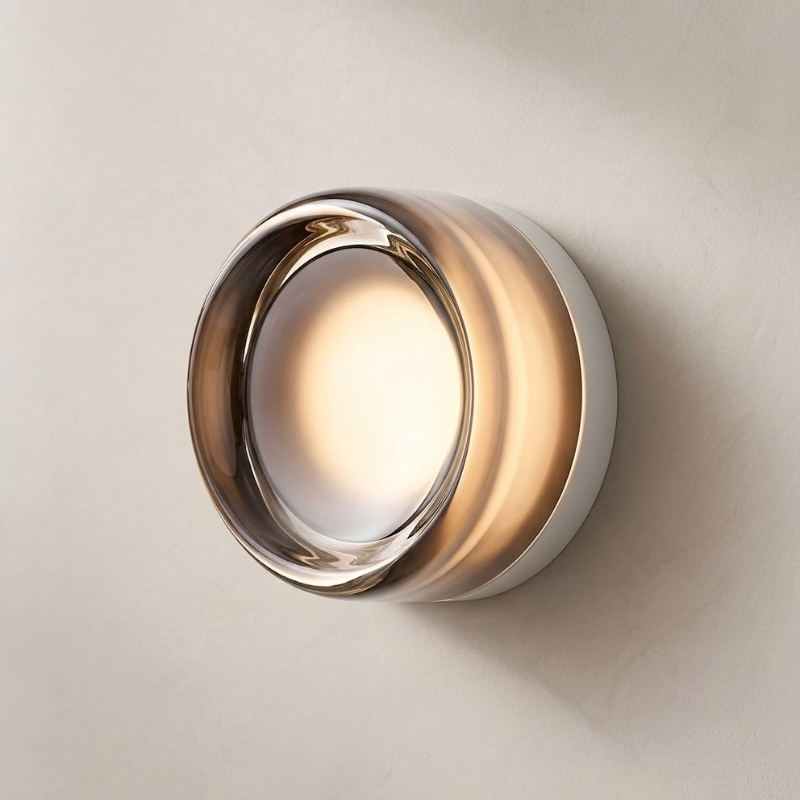 A modern cylindrical wall sconce with a brushed silver metallic body and a warm glowing light inside, mounted on a textured beige wall.
