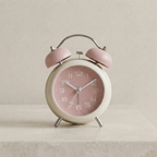A retro-style twin-bell alarm clock with a dusty pink face and matching pink bells, set on a light beige stone surface against a neutral beige background.