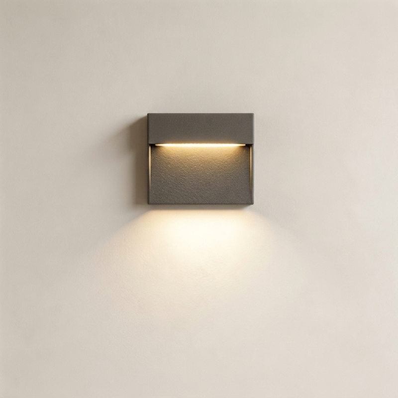 A modern square wall-mounted light fixture with a dark gray textured finish, emitting a warm yellow light downward against a plain off-white wall.