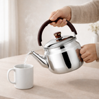 Person pouring water from a silver teapot into a white mug on a light surface.