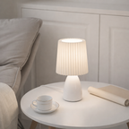 White table lamp on a round white table with a cup and saucer in a bedroom setting.