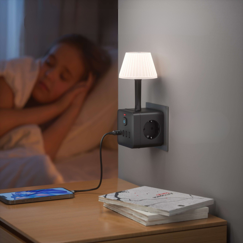 A wall-mounted power strip with a small lamp attached, plugged into a bedside outlet. A person is blurred in bed in the background, with a smartphone and magazines on the bedside table in the foreground.