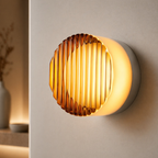 A modern wall-mounted light fixture with a ribbed amber glass shade and a glowing warm white cylindrical body, shown mounted on a beige wall in an interior setting.
