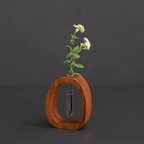 A small wooden oval-shaped vase with a clear glass tube in the center holding a delicate green plant with white flowers, set against a dark gray background.