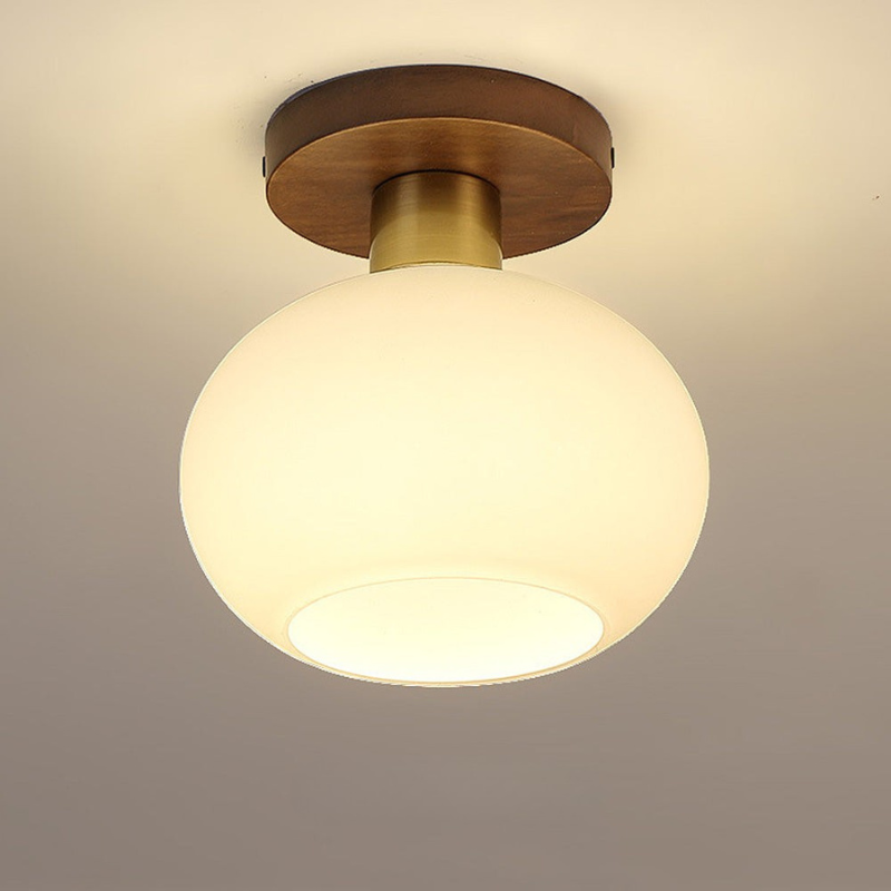A modern semi-flush mount ceiling light featuring a large, glowing frosted glass globe shade and a brushed brass and dark wood ceiling mount, shown installed on a neutral gray ceiling.