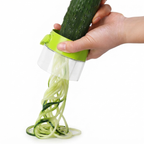 A hand holding a cucumber over a green and clear spiralizer, with pale green zucchini noodles being extruded against a white background.