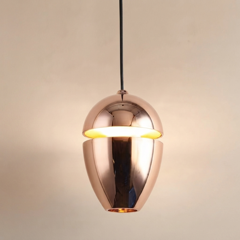 A modern pendant light with a metallic rose gold finish, featuring a split upper dome and a curved lower shade, illuminated with a warm yellow glow against a neutral beige background.