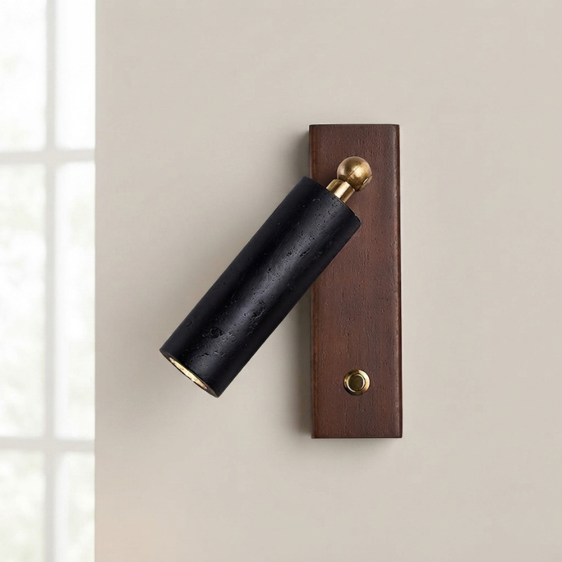 A modern wall-mounted spotlight with a matte black cylindrical shade, a dark brown rectangular backplate, and brass-toned hardware, shown against a warm beige wall.