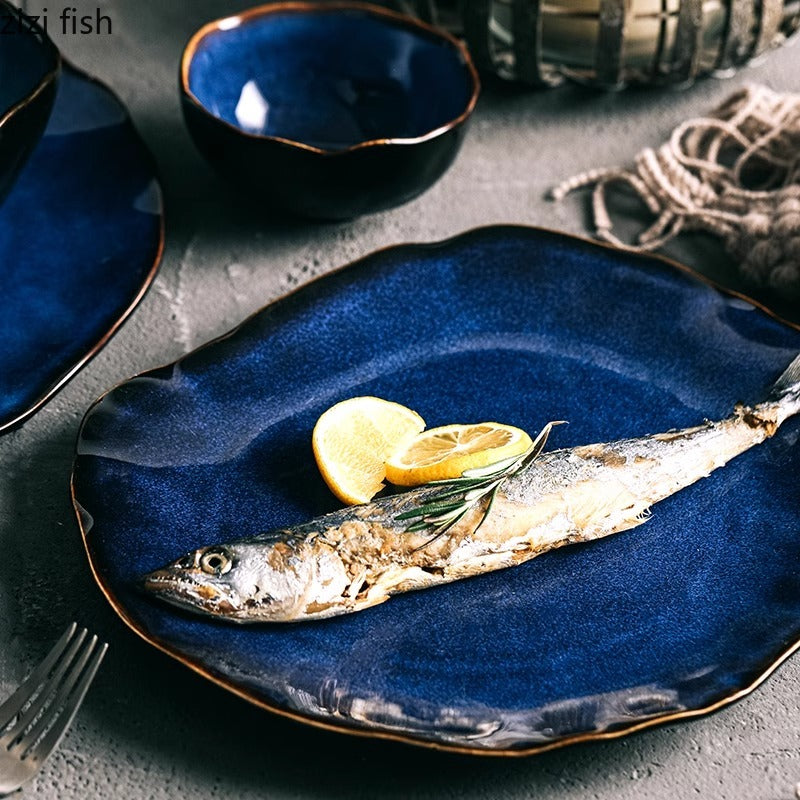 A dark blue ceramic plate with a gold rim, styled with a cooked fish, lemon slices, and a sprig of rosemary on a gray textured tabletop. Other matching blue dishes and tableware are partially visible in the background.