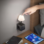 A bedside wall-mounted power strip with a small pleated lamp attached, shown in a bedroom setting. A person's hand is touching the lamp switch, and a tablet, notebook, and bed are visible nearby.