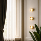 Three round wall sconces with warm yellow light are mounted vertically on a cream paneled wall next to a window with sheer curtains and a green plant in the foreground.