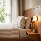 A neatly made bed with beige and white striped bedding and matching pillows in a modern bedroom. A wooden floating bedside table holds a lit wall lamp, a clear glass bottle, and a small metal cup.