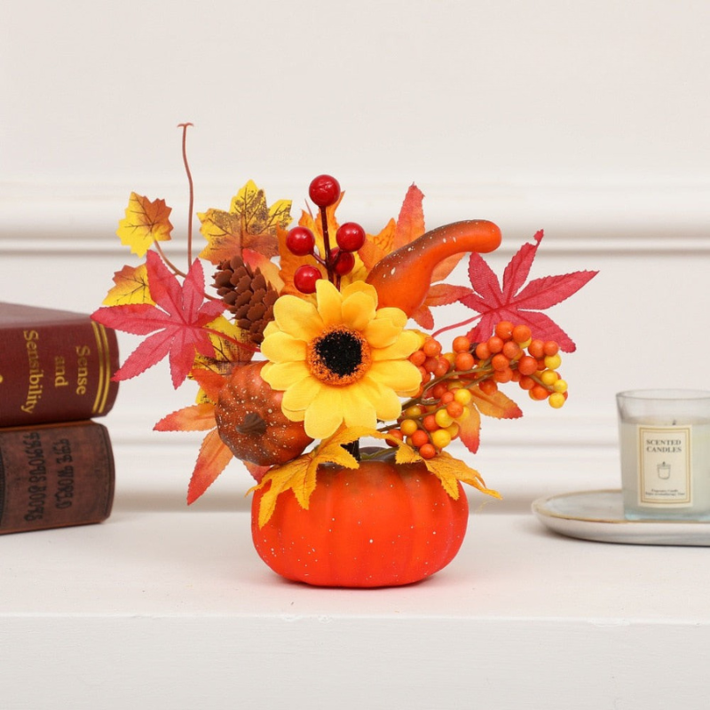 Autumn floral arrangement in a small orange pumpkin-shaped vase, featuring a yellow sunflower, red maple leaves, orange berries, pinecones, and faux gourds, styled on a white surface with books and a candle in the background.