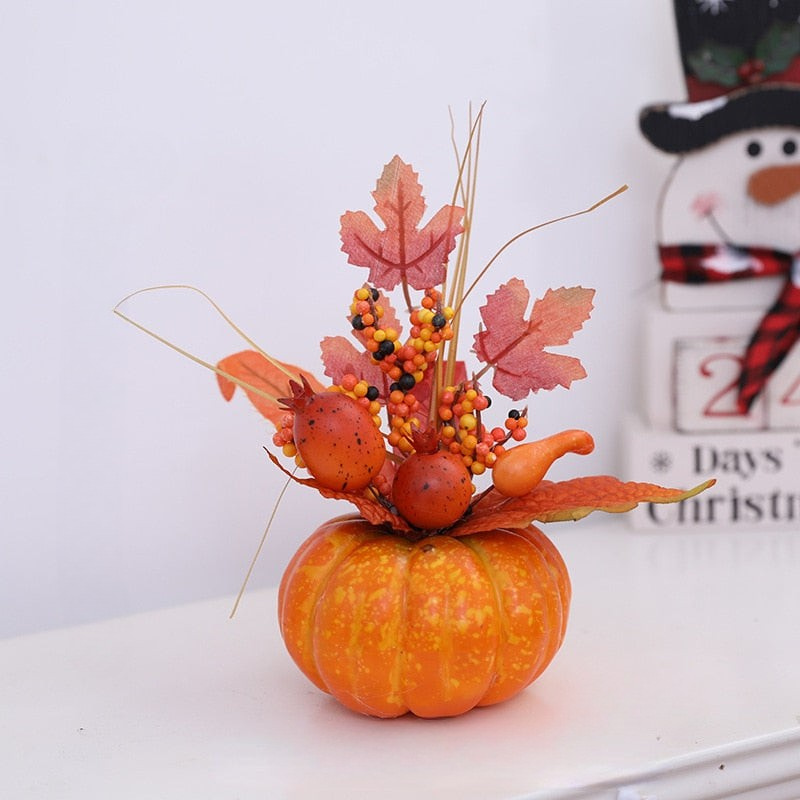 An autumn-themed decorative centerpiece featuring a small orange pumpkin base with faux maple leaves, berries, and dried grass stems, styled on a white surface with a snowman decoration in the background.