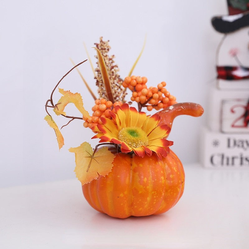 An autumn floral arrangement in a small orange pumpkin vase, featuring a large yellow and red flower, orange berries, tan grasses, and fall leaves, set on a white surface with a blurred holiday background.