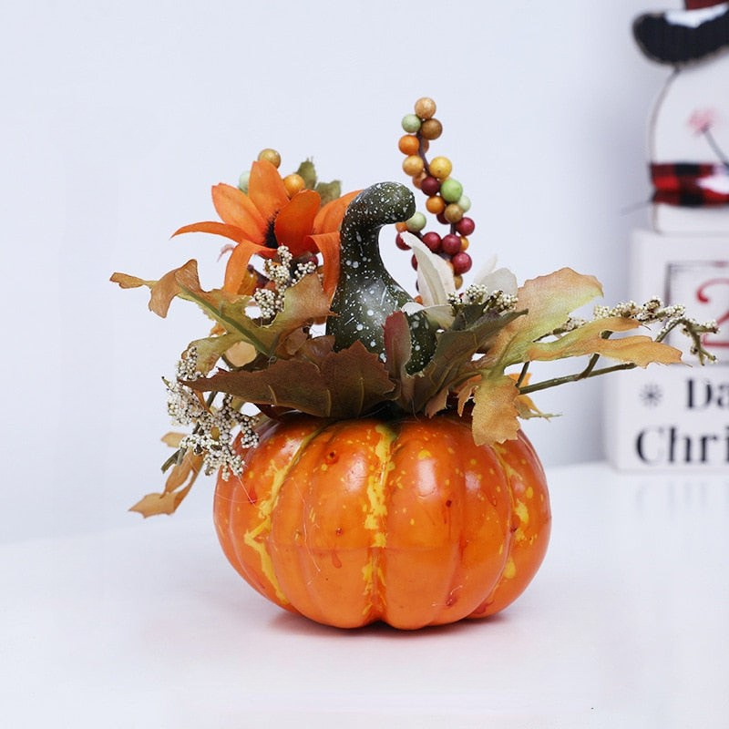 An autumn-themed decorative centerpiece featuring a small orange pumpkin base filled with faux fall leaves, an orange flower, a dark green bird figurine, and a string of multicolored berries, set against a clean white background.