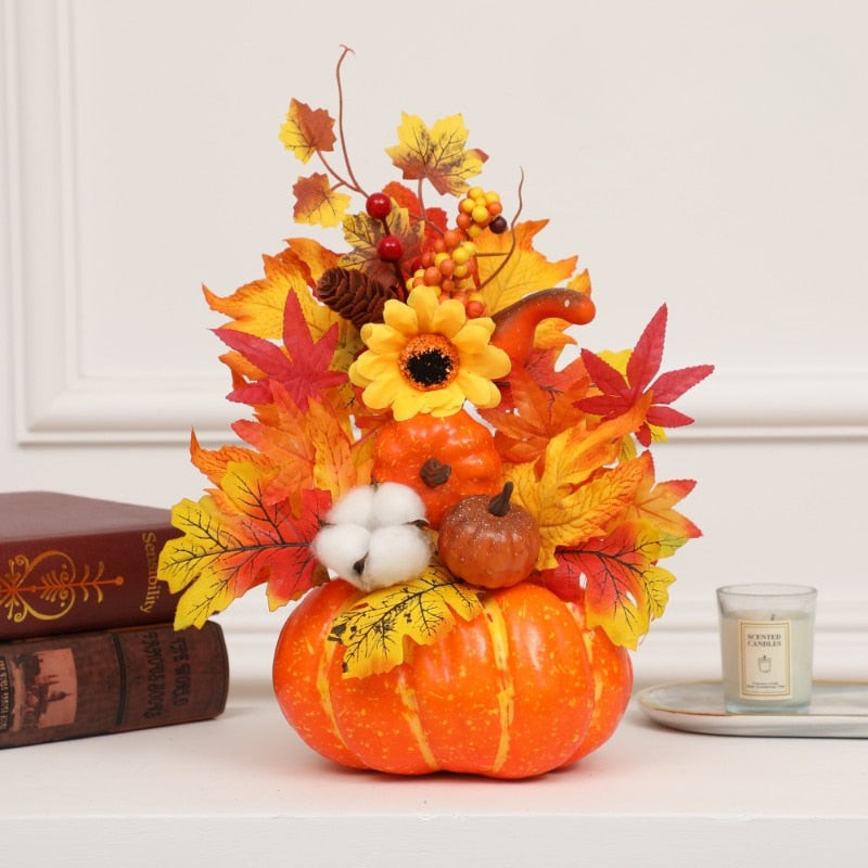 An autumn-themed decorative centerpiece featuring a small orange pumpkin base filled with faux fall leaves in red, orange, and yellow, a yellow sunflower, cotton boll, pinecone, and berries, styled on a white surface with books and a candle in the background.