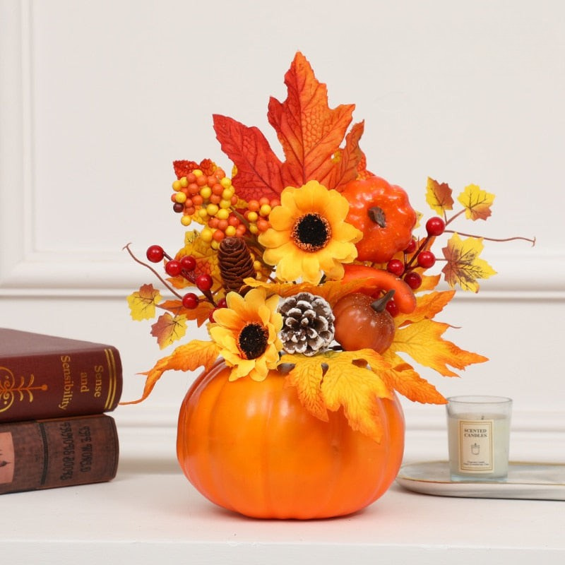An autumn floral arrangement in a bright orange pumpkin-shaped vase, featuring yellow sunflowers, red berries, orange maple leaves, pinecones, and small pumpkins, styled on a white surface with books and a candle.