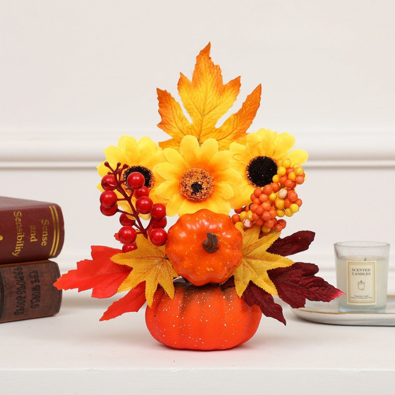 An autumn-themed floral arrangement in a small orange pumpkin-shaped vase, featuring yellow sunflowers, orange maple leaves, red berries, and a miniature orange pumpkin, styled on a white shelf with books and a candle.