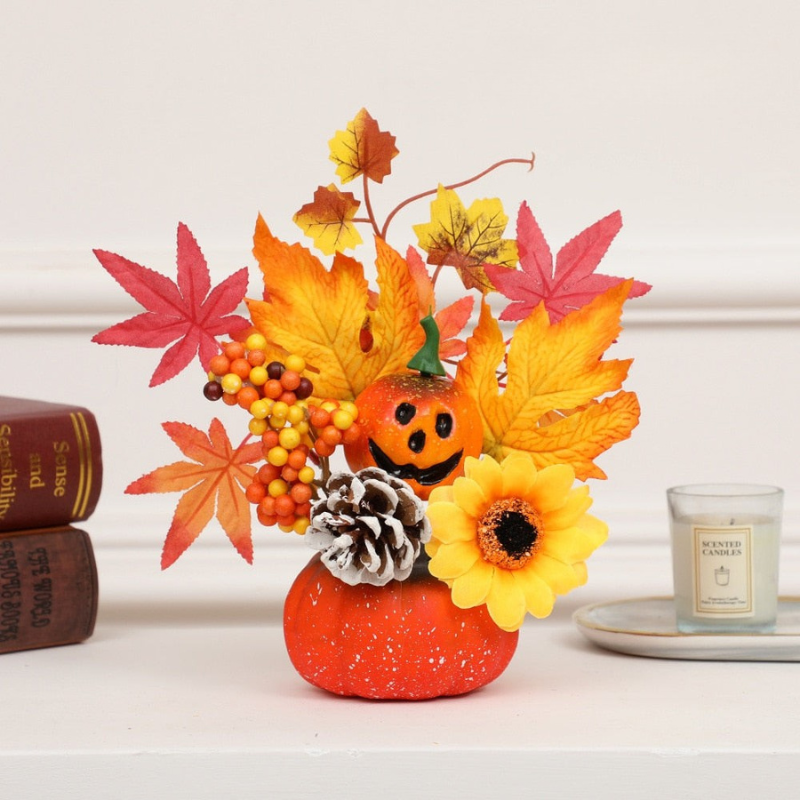 An autumn-themed decorative arrangement in a small orange pumpkin-shaped vase, featuring colorful fall leaves, a carved jack-o'-lantern, yellow sunflower, pinecone, and berries, styled on a white surface with books and a candle in the background.