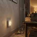 A square white wall light with four glowing warm bulbs mounted on a beige wall in a cozy indoor hallway scene with a table, books, and a cat in the background.
