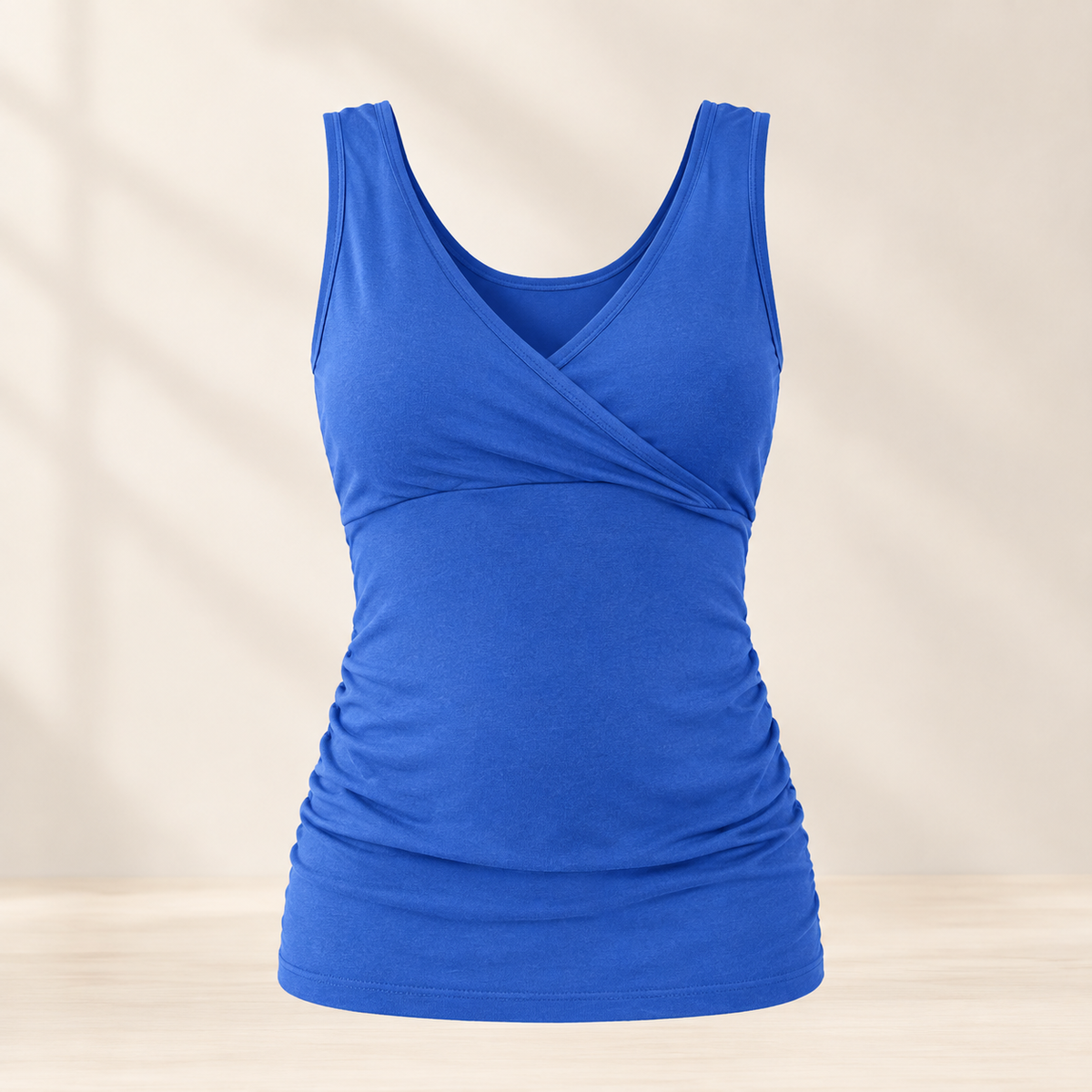 A royal blue sleeveless tank top with a crossover V-neckline and ruched waist details, displayed on a light beige background with soft shadow overlays.