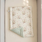 A baby quilted blanket with a cream background, featuring a pattern of pink rabbits in floral wreaths and small blue and pink dots, with a sage green minky dot reverse side, displayed on a beige mattress.