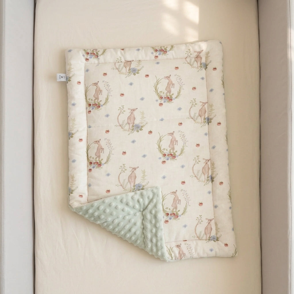 A baby quilted blanket with a cream background, featuring a pattern of pink rabbits in floral wreaths and small blue and pink dots, with a sage green minky dot reverse side, displayed on a beige mattress.