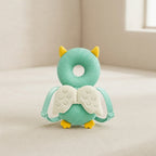 A mint green textured baby teether shaped like an owl, featuring yellow ears and feet, white wings, and adjustable straps, resting on a beige fabric surface.