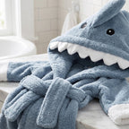 A plush blue shark-themed children's bathrobe with a hood shaped like a shark head, featuring white teeth and white cuffs, draped on a marble bathroom counter.