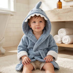 A toddler sitting on a rug in a bathroom, wearing a plush light blue hooded bathrobe designed like a shark with white trim and a white toothy jaw detail on the hood.