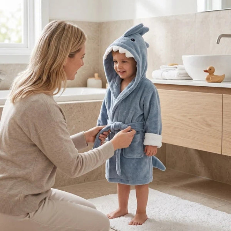 A child wearing a blue shark-shaped plush bathrobe stands in a bathroom while a woman adjusts the robe's tie. The bathroom has beige stone walls, a wooden vanity, a white sink, and a small rubber duck on the counter.