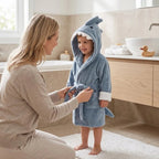 A child wearing a blue shark-shaped plush bathrobe stands in a bathroom while a woman adjusts the robe's tie. The bathroom has beige stone walls, a wooden vanity, a white sink, and a small rubber duck on the counter.