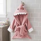 A pink plush shark-themed hooded bathrobe hanging on a wall hook in a bathroom setting. The robe features a shark head hood with embroidered eyes, white teeth, a tie belt, and white cuffs.