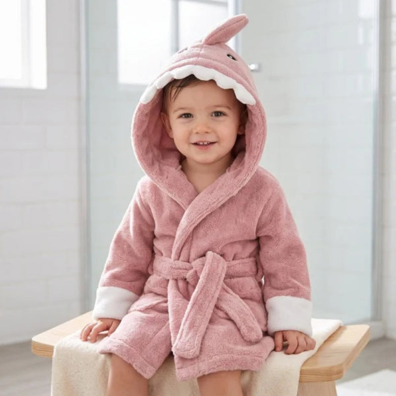 A smiling toddler wearing a plush pink hooded bathrobe with shark features, sitting on a wooden stool in a bright bathroom.