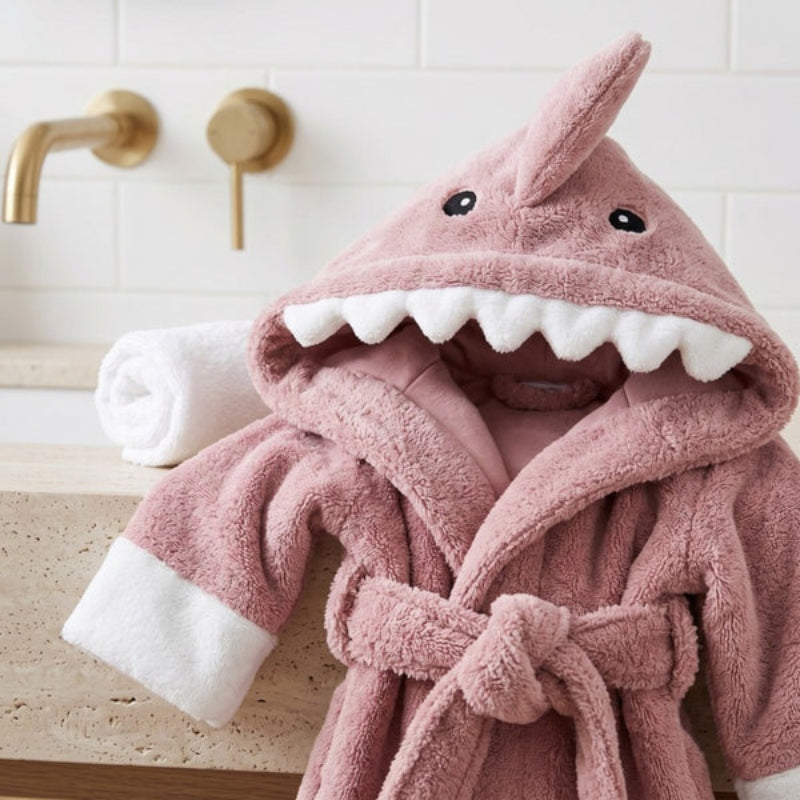 A pink plush hooded bathrobe designed like a shark, with white faux-teeth along the mouth, black eyes, and white cuffs, styled on a bathroom counter next to a rolled white towel.