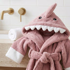 A pink plush hooded bathrobe designed like a shark, with white faux-teeth along the mouth, black eyes, and white cuffs, styled on a bathroom counter next to a rolled white towel.