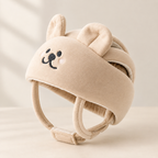 A beige baby helmet with plush bear ears and a cute embroidered bear face on the front, shown on a soft cream background.