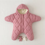 A pink quilted baby snowsuit with a hood and foot coverings, laid flat on a light textured background. The inside hood lining is cream-colored, and there are two small toys placed below the feet: a pink teddy bear and a white star.