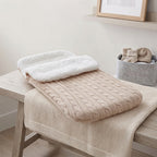 A beige cable-knit baby blanket with a white plush insert is draped over a light wood changing table in a softly styled nursery setting.