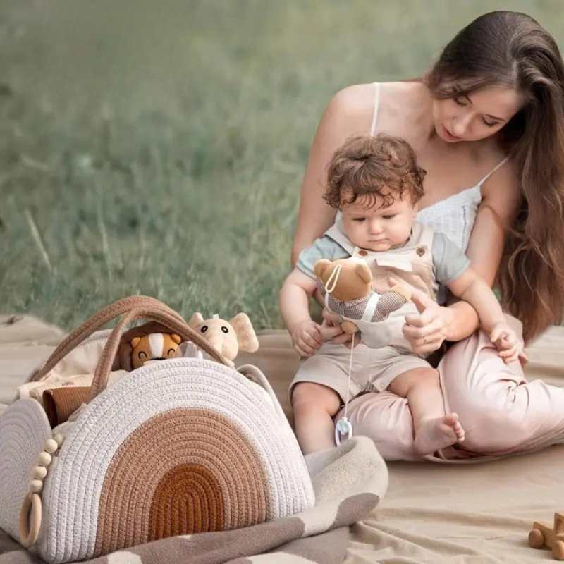 A woman and a baby sit on a blanket in a grassy outdoor setting, holding a plush toy, with a large woven rainbow-shaped baby bag in the foreground.