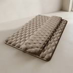 A rectangular bath mat with a raised tufted pattern, shown partially folded to display both sides. The mat is a warm taupe-beige color and is photographed on a matching neutral floor against a light beige wall.